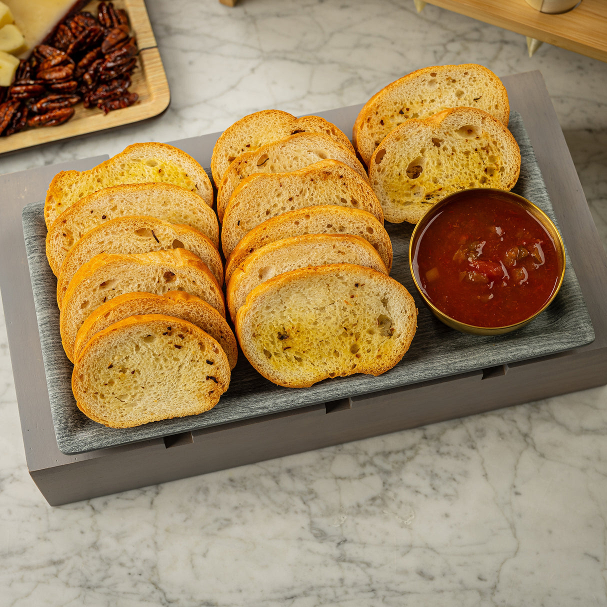 A rectangular melamine tray from TableCraft Products holds several slices of toasted baguette bread arranged neatly beside a bowl of chunky red sauce. The tray sits on a marbled surface, with pecans and cheese partially visible in the background.