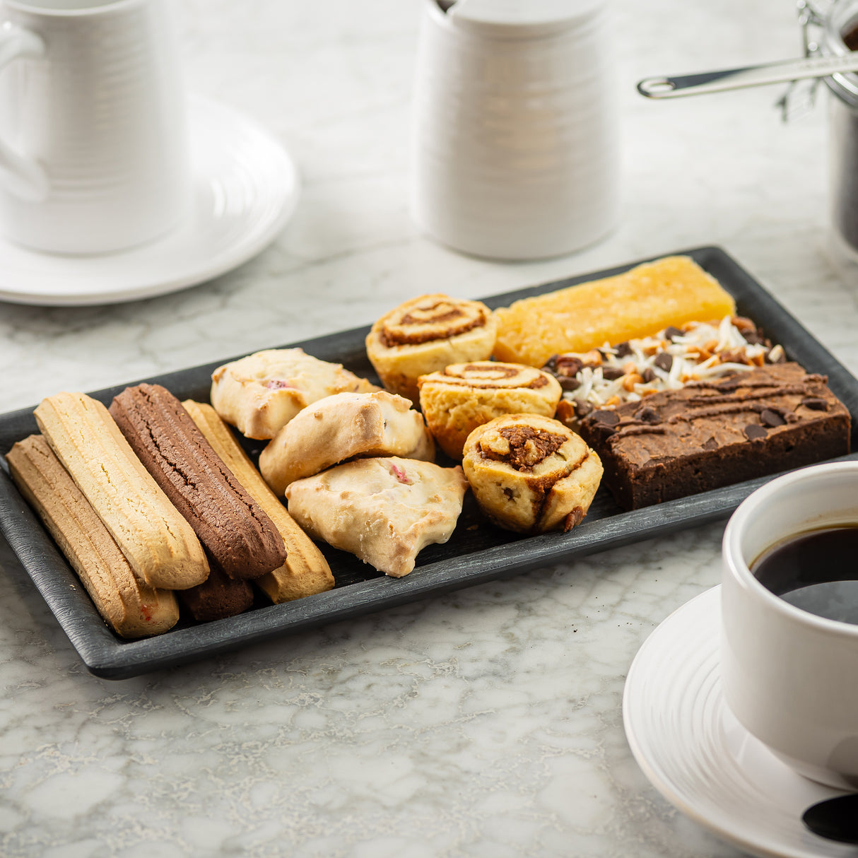 A rectangular serving tray filled with assorted pastries and cookies, including biscotti, brownies, and cinnamon rolls, sits on a marble table alongside a white cup of black coffee and ceramic dishes, creating a cozy café atmosphere.