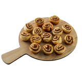 A round Serving Paddle from the Timber Melamine Collection holds fifteen small, spiral-shaped cinnamon rolls arranged neatly. The rolls have a golden-brown crust with visible cinnamon swirls. The background is plain white.