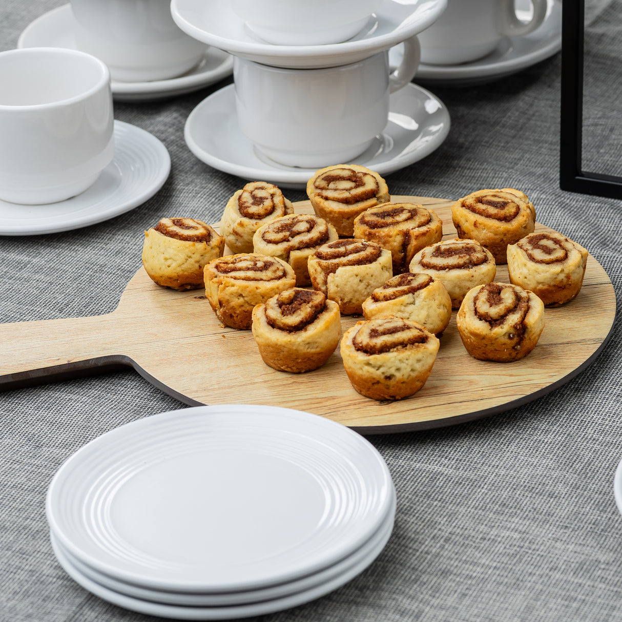 A wooden Serving Paddle with small cinnamon rolls rests on a gray tablecloth. Stacks of white plates and teacups with saucers add to the inviting setting, perfect for tea or coffee from the Timber Melamine Collection.