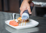 A hand squeezes barbecue sauce from a WideMouth Squeeze Bottle onto a sandwich on a white plate, with sliced tomatoes and fries beside it. The NSF-certified plate sits on a stainless steel surface in what appears to be a kitchen setting.