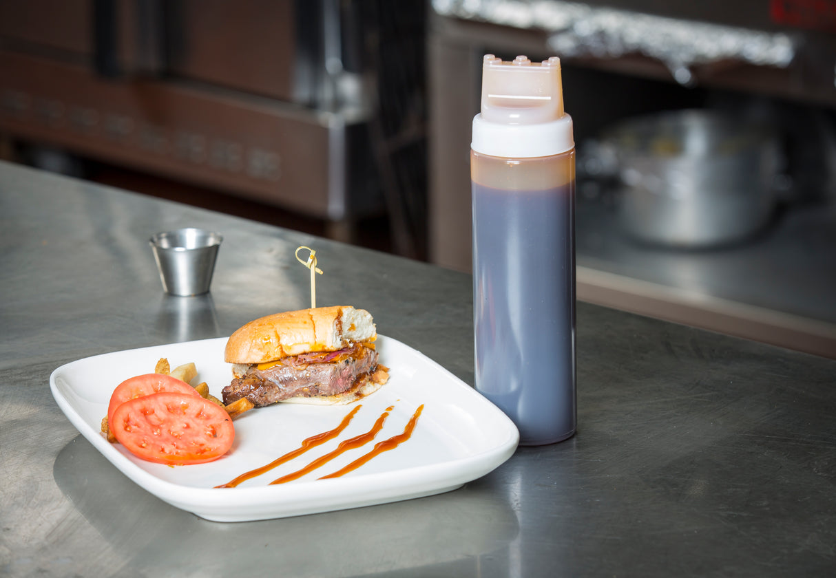 A sandwich with a bite taken out sits on a white plate, garnished with tomato slices and pickles. Nearby, an NSF-listed WideMouth Squeeze Bottle with Three Tip Top SelecTop stands on a stainless steel counter in a commercial kitchen.