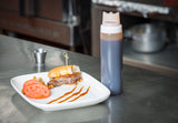 A sandwich with a bite taken out sits on a white plate, garnished with tomato slices and pickles. Nearby, an NSF-listed WideMouth Squeeze Bottle with Three Tip Top SelecTop stands on a stainless steel counter in a commercial kitchen.