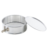 A round, stainless steel springform pan with a removable lid placed to the side. The pan has a metal latch on the left for secure closure, smooth shiny surfaces, and a snug-fitting lid matching its metallic finish.