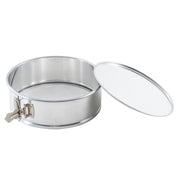 A round, stainless steel springform pan with a removable lid placed to the side. The pan has a metal latch on the left for secure closure, smooth shiny surfaces, and a snug-fitting lid matching its metallic finish.