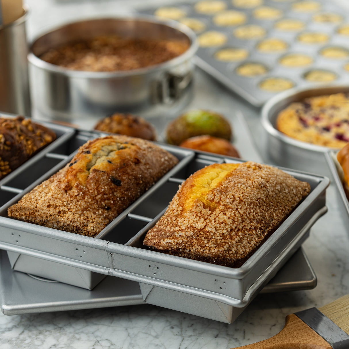 Two loaves of bread with golden, sugar-crusted tops sit in an aluminized steel bread pan. Behind them, muffins and cakes rest on a marble countertop, creating a warm, inviting baking scene filled with delicious homemade treats.
