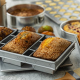 Two loaves of bread with golden, sugar-crusted tops sit in an aluminized steel bread pan. Behind them, muffins and cakes rest on a marble countertop, creating a warm, inviting baking scene filled with delicious homemade treats.