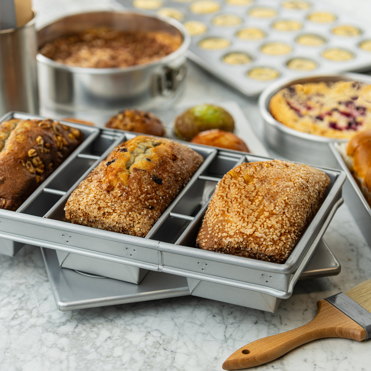 Three small, golden-brown loaves with textured, sugared crusts sit in a 3-loaf strapped bread pan on a marble counter. Around them are more baked goods, baking pans, and a pastry brush, evoking a busy, fresh-baked kitchen scene.