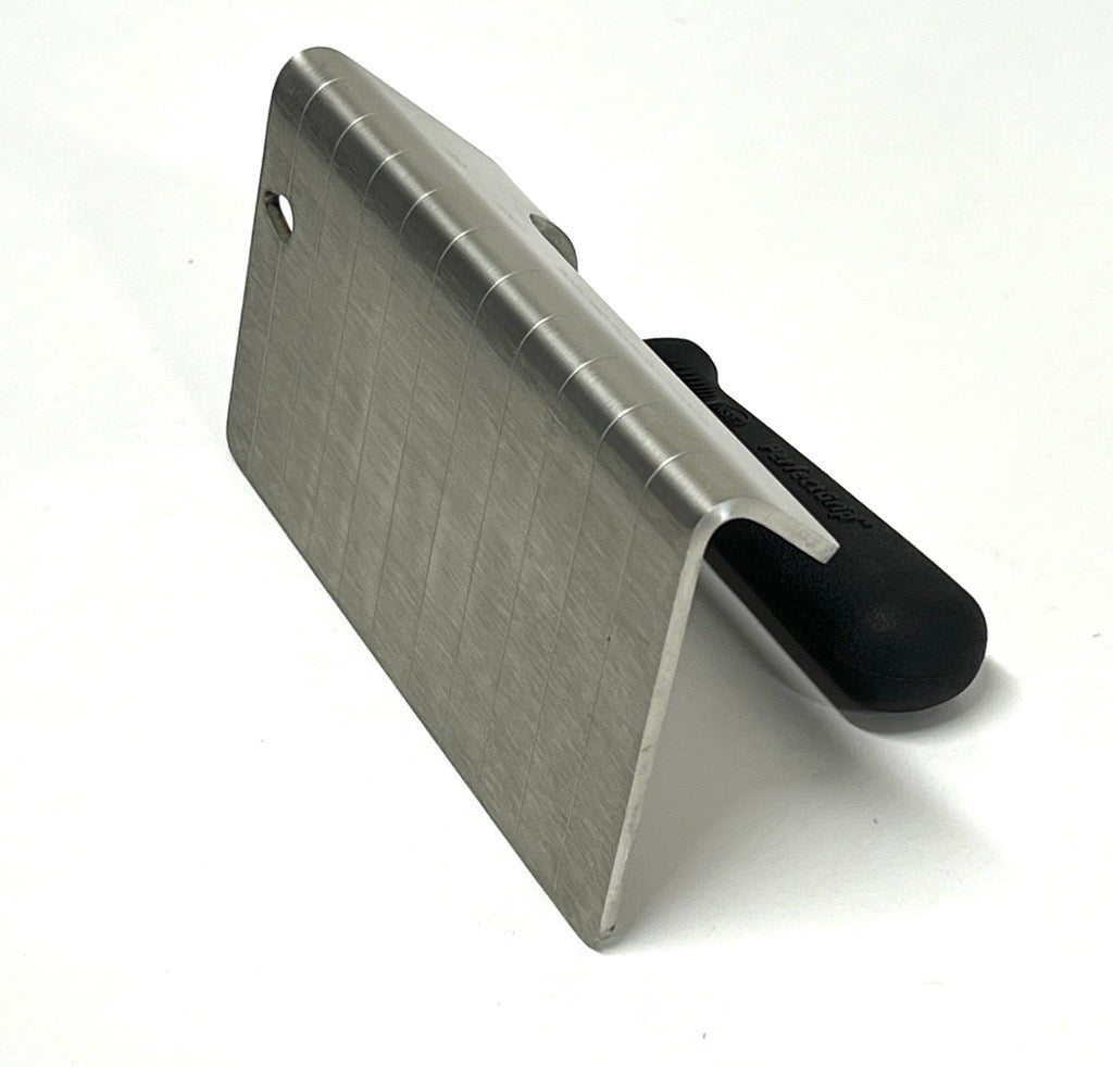 A stainless steel steak weight with NSF certification, this PerfectGrip Steak Weight features a wide, flat, L-shaped blade with horizontal grooves and a black handle at a right angle, resting on a white surface. A small hole sits near the top edge of the blade.