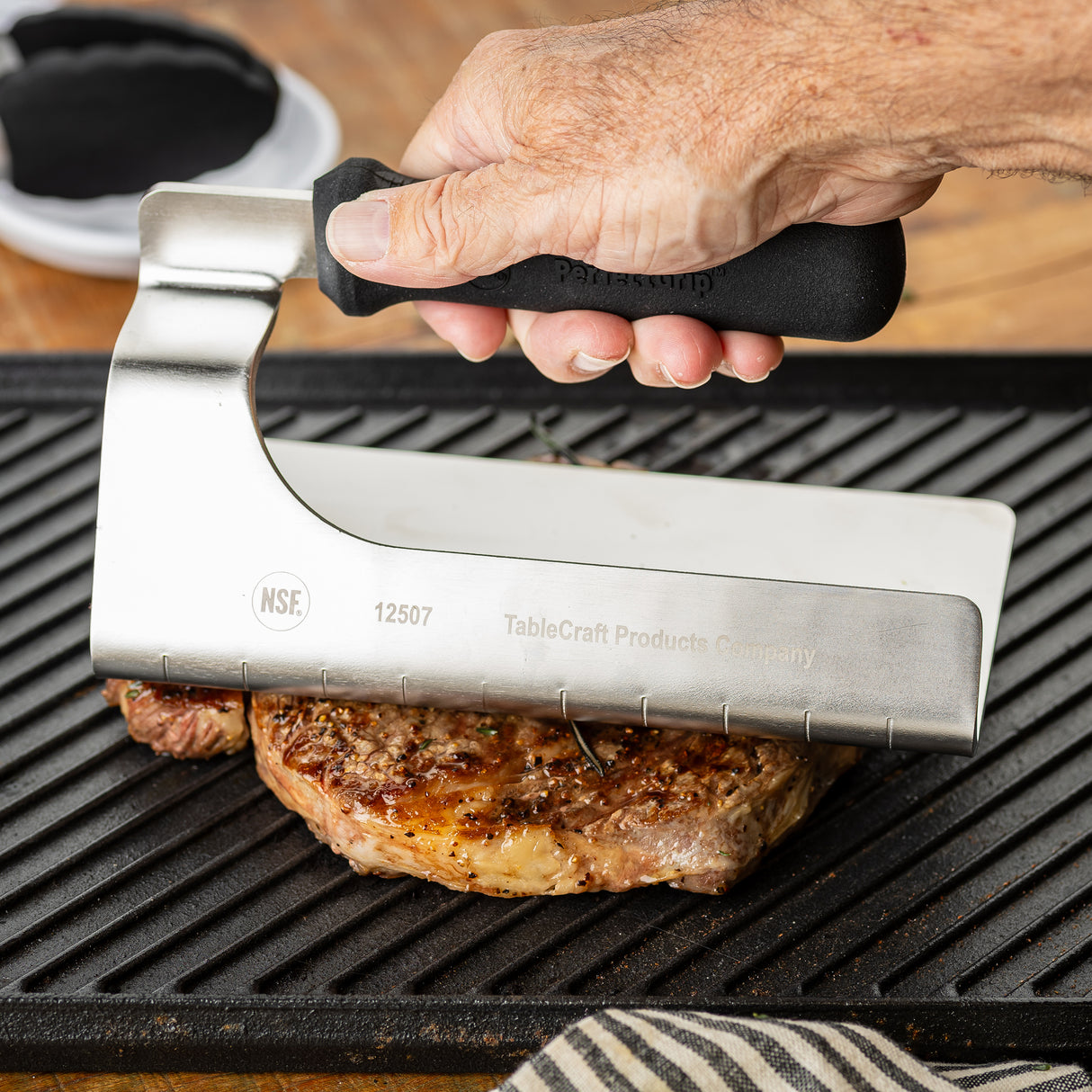 A hand presses the NSF-certified PerfectGrip Steak Weight, a stainless steel steak weight with a black handle, onto a juicy, well-seasoned steak on a ridged grill pan. The press features engraved marks and the brand name TableCraft Products Company.