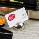 A white business card for TableCraft is held by a shiny, stainless steel card holder with a silver hammered pattern on a textured beige surface. The card displays a red logo and contact details for Sue Denice, Director of Sales. Food trays are partially visible in the background.