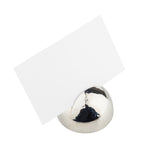 A blank white card is held upright by a silver hammered pattern card holder—a smooth, shiny sphere of stainless steel—against a plain white background. The reflective surface displays light and subtle reflections.
