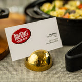 A business card in a gold stainless steel card holder with a hammered pattern displays the TableCraft logo and Sue Denica's contact info. In the background, a blurred dish hints at a restaurant or catering setting.
