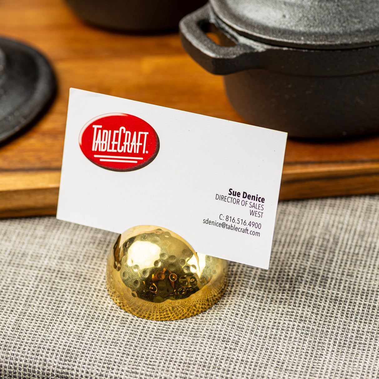 A white business card for Sue Denice, Director of Sales at TableCraft, sits in a gold stainless steel card holder with a hammered pattern on a gray textured cloth. In the background, black cast iron cookware rests on a wooden surface.