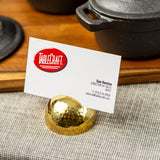 A white business card for Sue Denice, Director of Sales at TableCraft, sits in a gold stainless steel card holder with a hammered pattern on a gray textured cloth. In the background, black cast iron cookware rests on a wooden surface.