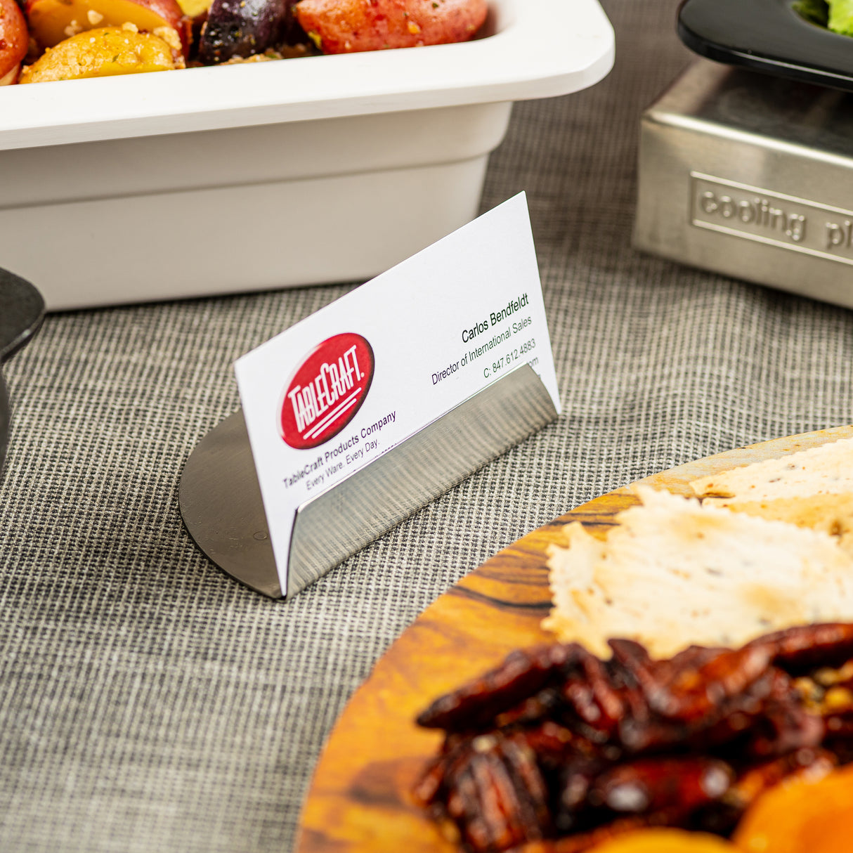 A close-up of a buffet table features a TableCraft stainless steel card holder displaying a business card for Carlos Benedetti. Surrounding the card are dishes with mixed vegetables and a wooden platter with crackers and pecans.