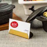A TableCraft business card for Sue Denice, Director of Sales, West, rests in a gold coated stainless steel Half Moon Card Holder on a grey textured tablecloth, surrounded by black cast iron pots with noodles inside.