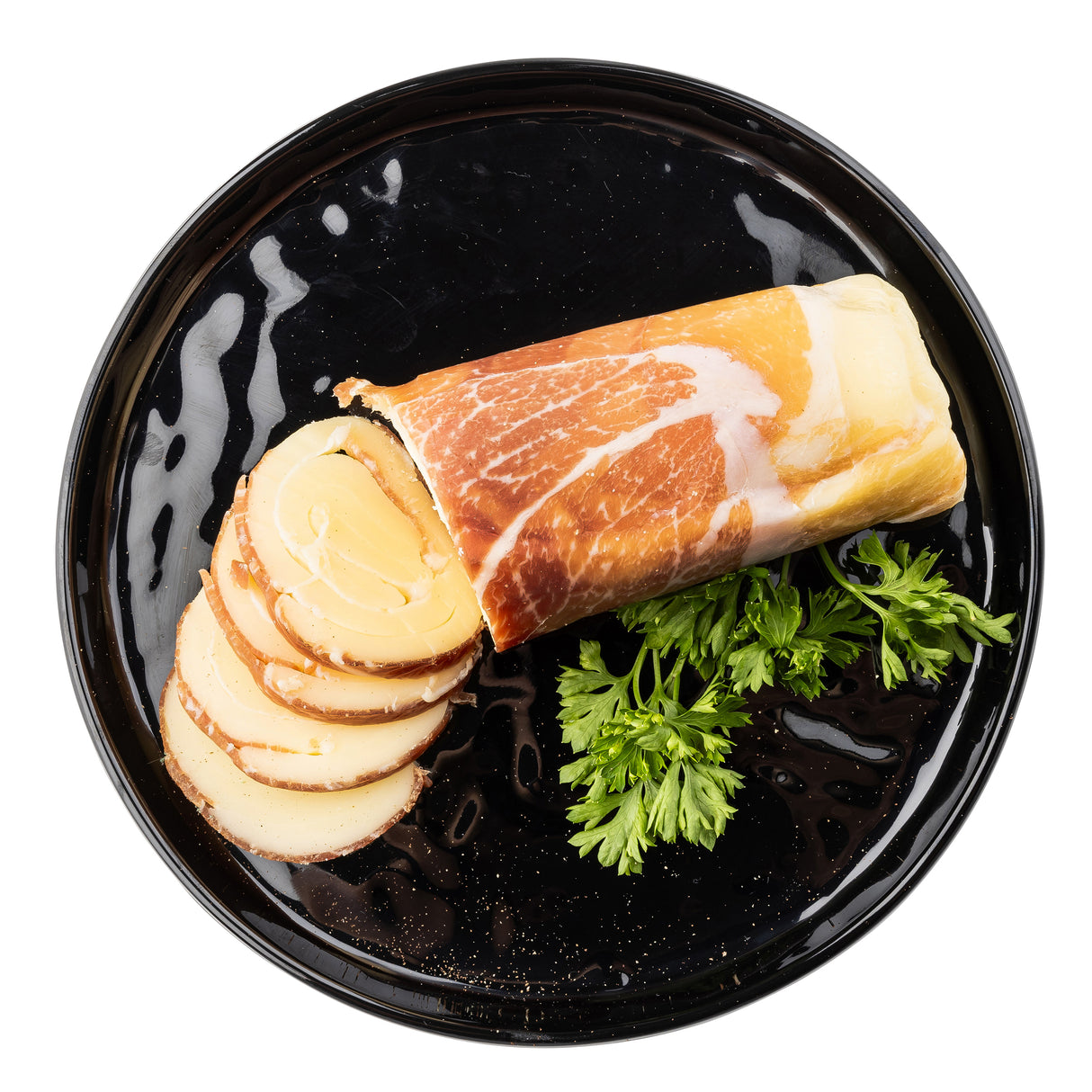 On a round black melamine serving plate by TableCraft, slices of rolled smoked cheese wrapped in prosciutto are arranged beside curly fresh parsley. The partially sliced cheese reveals a spiral pattern inside against a white background.