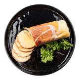 On a round black melamine serving plate by TableCraft, slices of rolled smoked cheese wrapped in prosciutto are arranged beside curly fresh parsley. The partially sliced cheese reveals a spiral pattern inside against a white background.