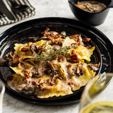A Ridge Collection Serving Plate holds ravioli topped with creamy mushroom sauce, sautéed mushrooms, herbs, and bacon bits on a marble table. A bowl of extra mushroom sauce and utensils are in the background. Fresh thyme garnishes the dish.