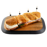 A long sandwich with ham, salami, provolone cheese, and spinach on a soft bun sits on a TableCraft Acacia Collection serving board with a black border, cut into sections and secured with toothpicks topped with black olives.