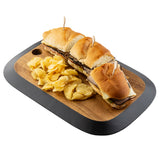A TableCraft Acacia Collection rectangular serving board holds three mini roast beef sandwiches with cheese, each secured with a toothpick, alongside a pile of ridged potato chips. The board features black trim and a circular cutout handle.