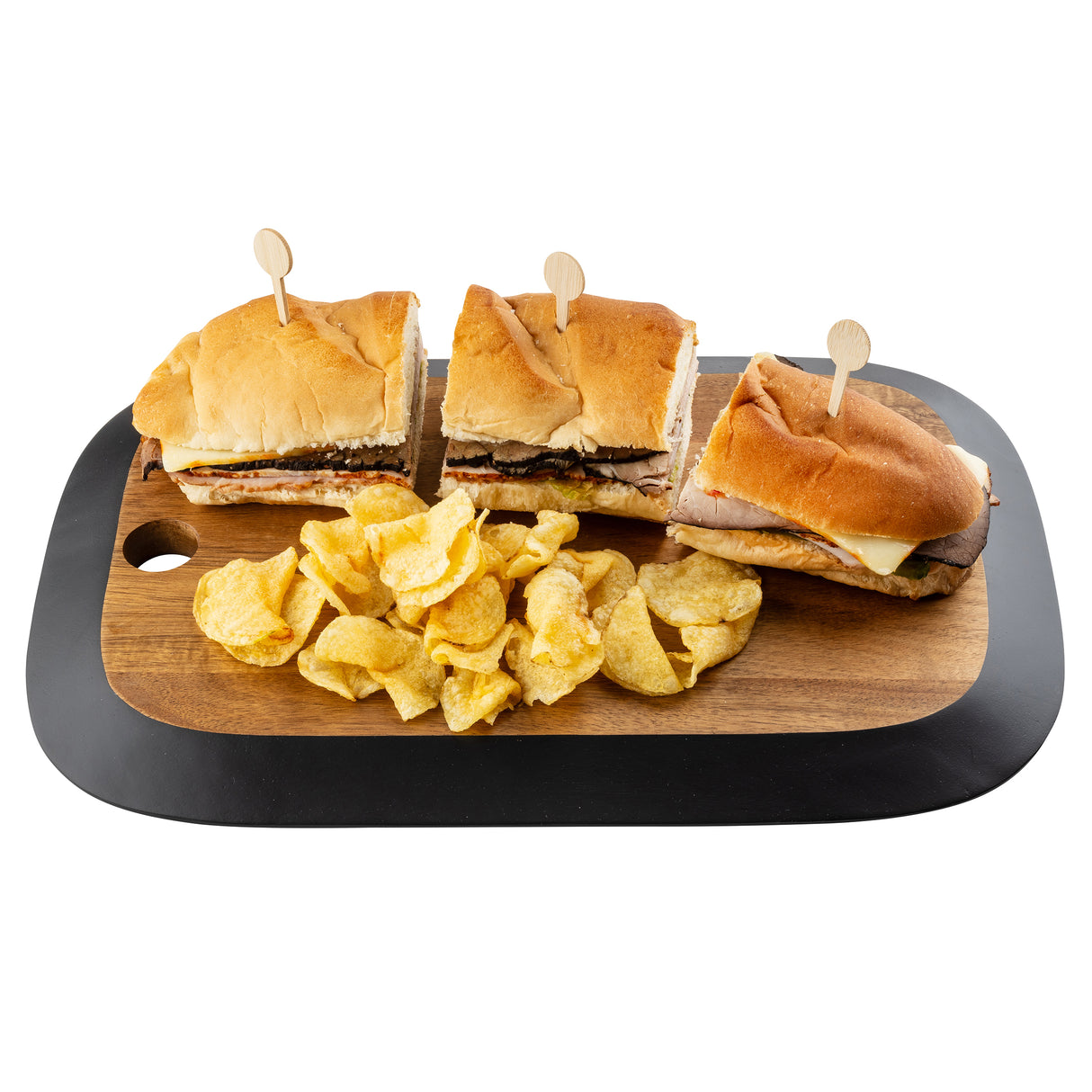 A TableCraft Acacia Collection rectangular serving board holds three sandwich halves, each with sliced meat and cheese, secured by toothpicks. Ridged potato chips are piled in front. The background is white.