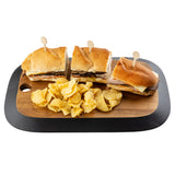 A TableCraft Acacia Collection rectangular serving board holds three sandwich halves, each with sliced meat and cheese, secured by toothpicks. Ridged potato chips are piled in front. The background is white.