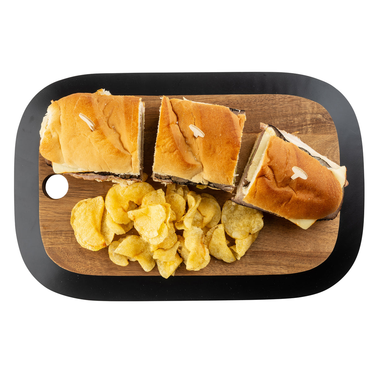 A TableCraft Products rectangular Acacia serving board holds three sandwich halves with golden buns and toothpicks, arranged in a row. Below them is a pile of crinkled potato chips. The board has a rounded black border and a cutout in the top left corner.