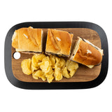 A TableCraft Products rectangular Acacia serving board holds three sandwich halves with golden buns and toothpicks, arranged in a row. Below them is a pile of crinkled potato chips. The board has a rounded black border and a cutout in the top left corner.