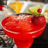 A close-up of a vibrant red frozen strawberry cocktail in a margarita glass, garnished with a fresh strawberry and a small red berry on TableCraft 12582 red cocktail picks. In the blurred background, sliced lemons and limes are visible in white trays.