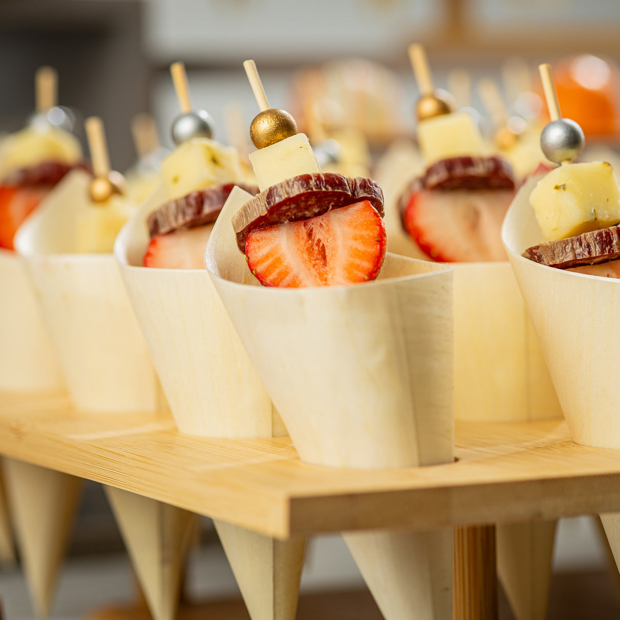 Several wooden cones are filled with skewers holding cubes of cheese, cured sausage slices, and halved strawberries. Each skewer, crafted with gold picks or bamboo ball picks like TableCraft 12583, is elegantly arranged in a wooden display stand.