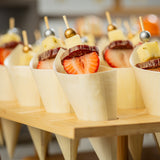 Several wooden cones are filled with skewers holding cubes of cheese, cured sausage slices, and halved strawberries. Each skewer, crafted with gold picks or bamboo ball picks like TableCraft 12583, is elegantly arranged in a wooden display stand.