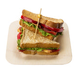 A sandwich cut in half is stacked on a light wooden plate. Filled with lettuce, tomato slices, crispy bacon, and toasted bread, it’s held together with elegant gold picks. The white background makes the sandwich and plate stand out clearly.