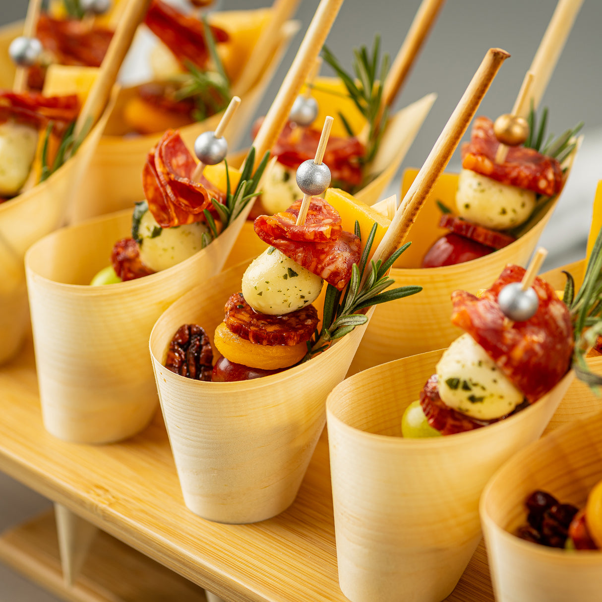 Individual bamboo cups from TableCraft, each with Ball Picks skewering marinated mozzarella, dried apricots, salami, and fresh rosemary. Pecans and green grapes complete the selection, all neatly arranged on a wooden tray for serving.