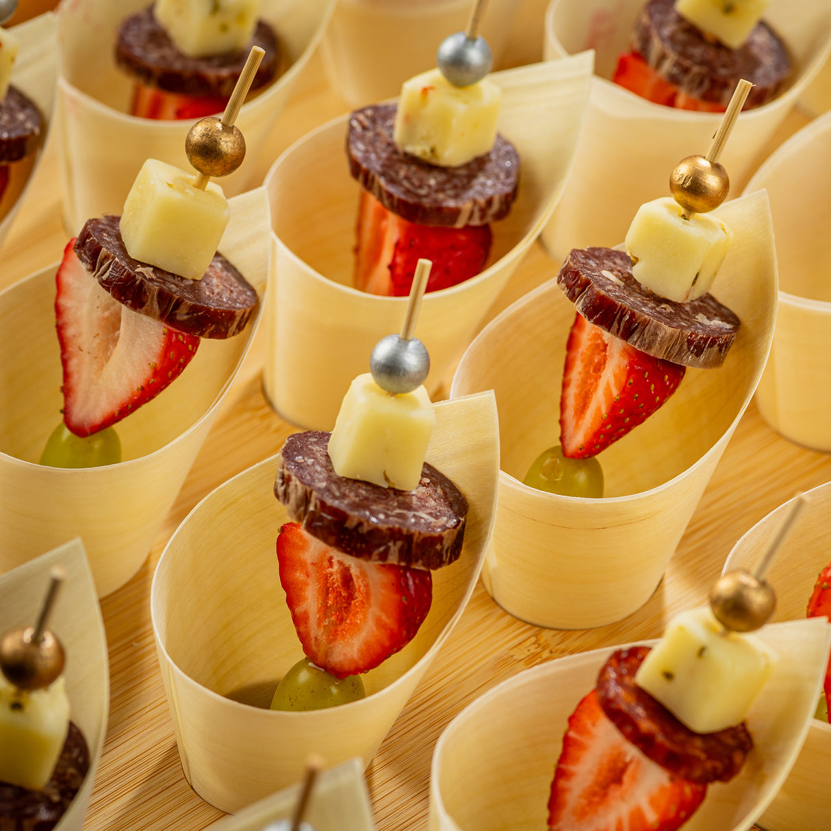Wooden cups each hold a Ball Pick skewer with a grape, strawberry slice, salami slice, and cheese cube. The bamboo skewers have metallic beads on top and are neatly arranged in rows on a light wooden surface.