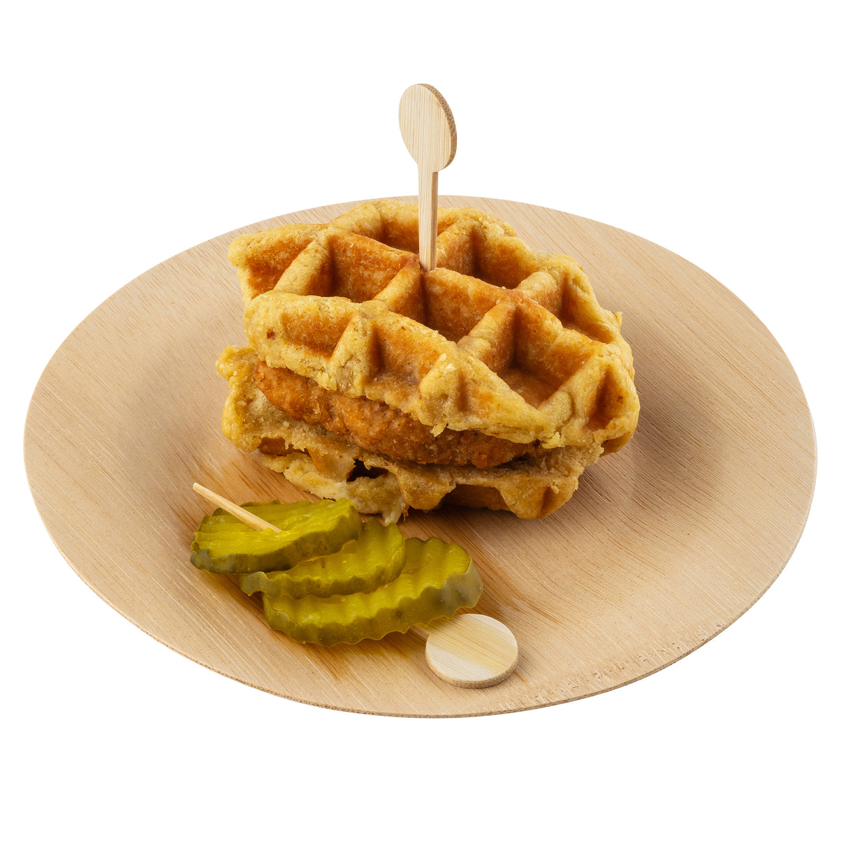 A sandwich made of two thick waffles with fried chicken inside, held together by a TableCraft bamboo pick, is served on a round bamboo plate. Three crinkle-cut pickle slices are placed beside the sandwich on the plate.