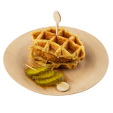 A sandwich made of two thick waffles with fried chicken inside, held together by a TableCraft bamboo pick, is served on a round bamboo plate. Three crinkle-cut pickle slices are placed beside the sandwich on the plate.