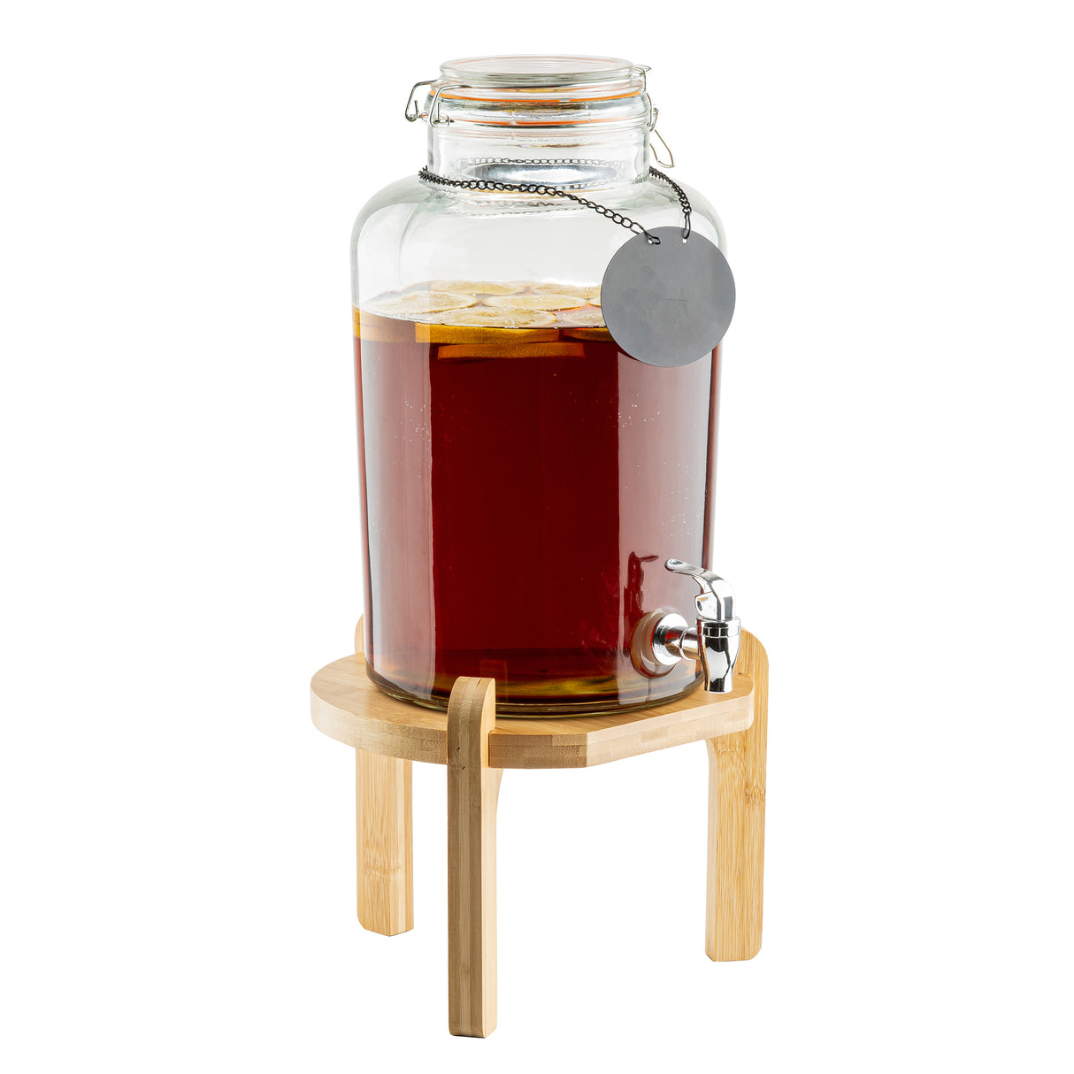 A large glass beverage dispenser filled with dark liquid and lemon slices sits on a TableCraft 12652 bamboo beverage dispenser stand. It features a metal spigot, a blank round tag on a chain, and a clear glass lid with a clasp.