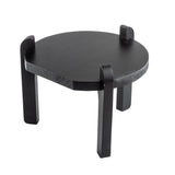 A small, round, black painted wood stand with three thick, rectangular legs that extend slightly above the tabletop. The modern, minimalist design features a smooth finish and low profile, ideal as a TableCraft stand against a white background.