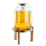 A large TableCraft glass beverage dispenser with a metal lid and silver spigot sits on an acacia wood stand. Filled with yellow-orange liquid, it features an ice core insert to keep drinks cold. The background is plain white.