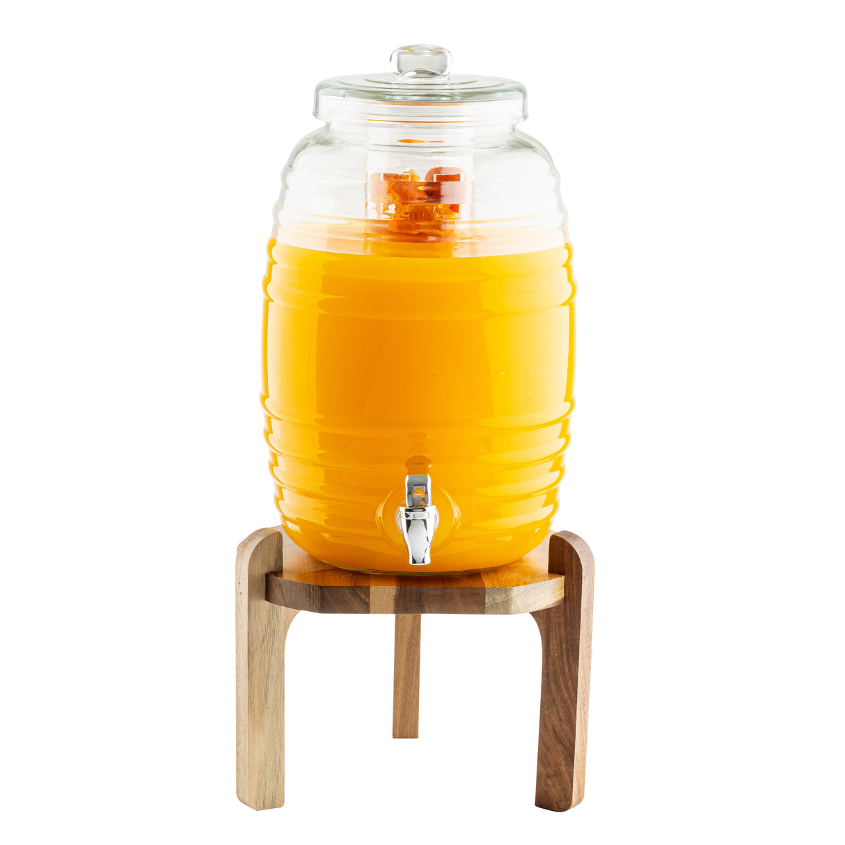 A TableCraft glass beverage dispenser filled with bright orange juice sits on an acacia wood stand. The dispenser has a lid, a metal spigot at the bottom, and visible orange slices inside. The background is plain white.