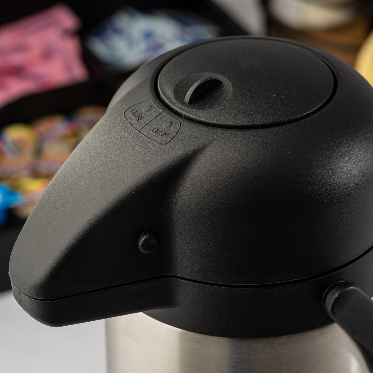 Close-up of a stainless steel Airpot coffee dispenser with a push-button lid labeled LOCK and UNLOCK. In the blurred background, packets of sugar and creamer are visible, slightly out of focus.