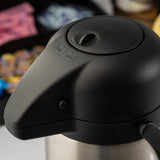 Close-up of a stainless steel Airpot coffee dispenser with a push-button lid labeled LOCK and UNLOCK. In the blurred background, packets of sugar and creamer are visible, slightly out of focus.