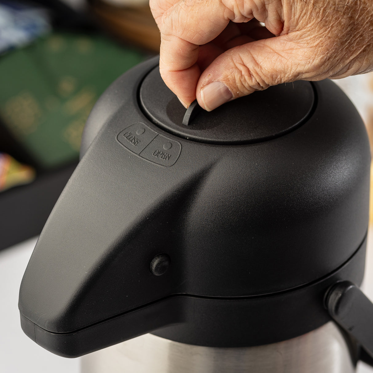 A close-up of a hand lifting the tab on the lid of a black and stainless steel vacuum insulated Airpot. The lid has “CLOSE” and “OPEN” markers next to the tab. The background is blurred, with green and colorful items partially visible.