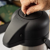 A close-up of a hand lifting the tab on the lid of a black and stainless steel vacuum insulated Airpot. The lid has “CLOSE” and “OPEN” markers next to the tab. The background is blurred, with green and colorful items partially visible.
