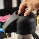 A close-up of a hand pressing the pump button on a black, stainless steel Airpot dispenser. The spout dispenses liquid, with labeled LOCK and UNLOCK buttons. Blurred background hints at colorful objects and a basket.