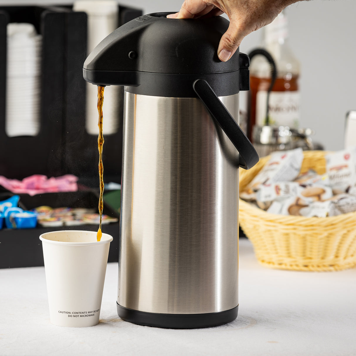 A hand presses the top of a vacuum insulated stainless steel airpot, dispensing coffee into a white paper cup. In the background are coffee supplies: sweeteners, creamers in a basket, and stacked cups on a light-colored countertop.