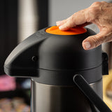 A close-up of a hand pressing an orange button on top of a black stainless steel airpot coffee dispenser, with a blurred background featuring cups and colorful objects. The scene suggests a beverage station or coffee setup.