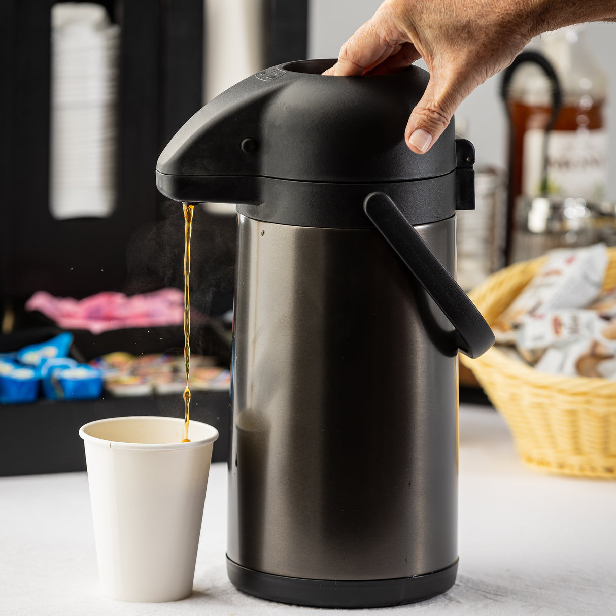 A hand presses down on the black lid of a stainless steel airpot, dispensing hot coffee into a white paper cup. In the background, vacuum insulated coffee supplies like sugar packets, creamers, and a basket with lids can be seen as steam rises from the cup.
